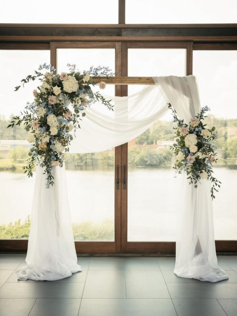Wooden wedding arch draped in white fabric with blue and white floral arrangements by a lake.