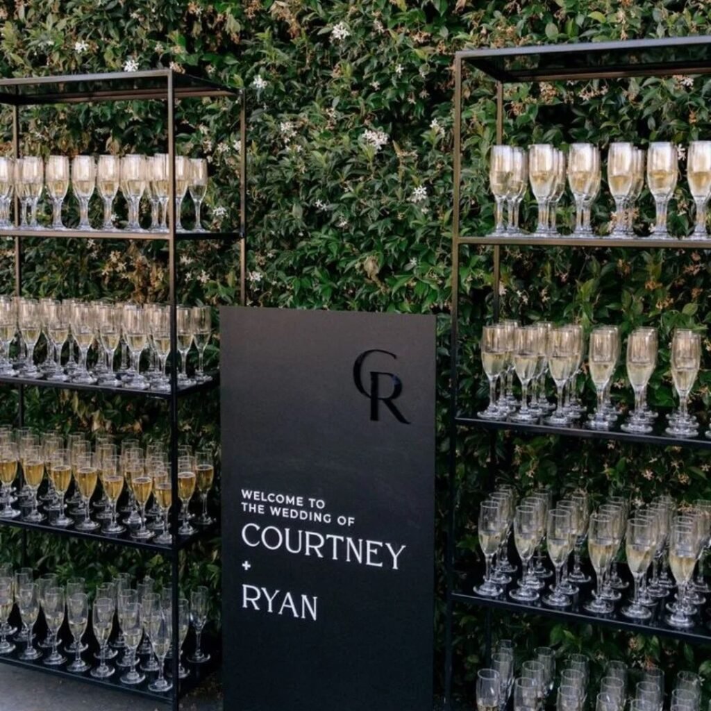 Champagne glasses on black metal shelves next to a wedding welcome sign against a green hedge.