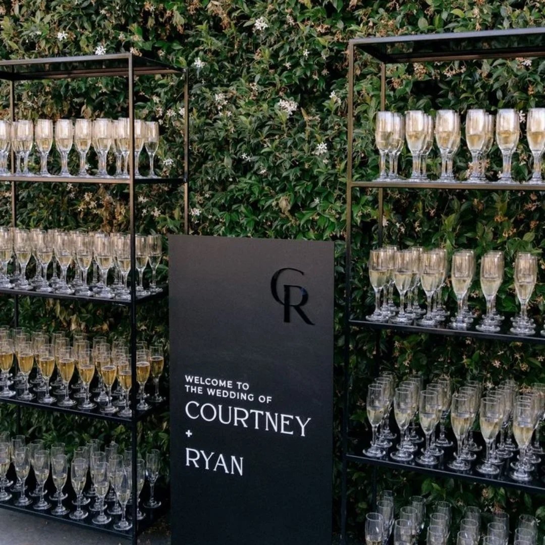Champagne glasses on black metal shelves next to a wedding welcome sign against a green hedge.