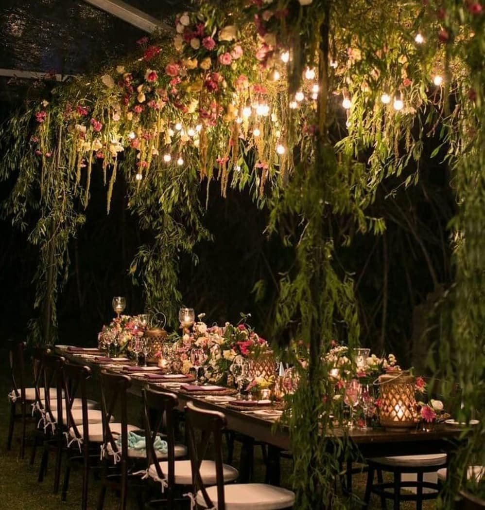 Long dining table decorated with abundant pink flowers and hanging greenery under glowing warm lights.
