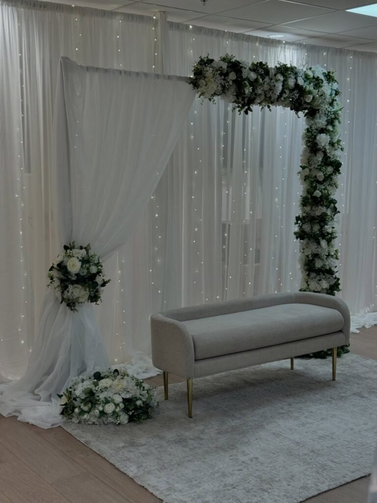 White floral arch and draped fabric backdrop with a grey chaise lounge on a rug.