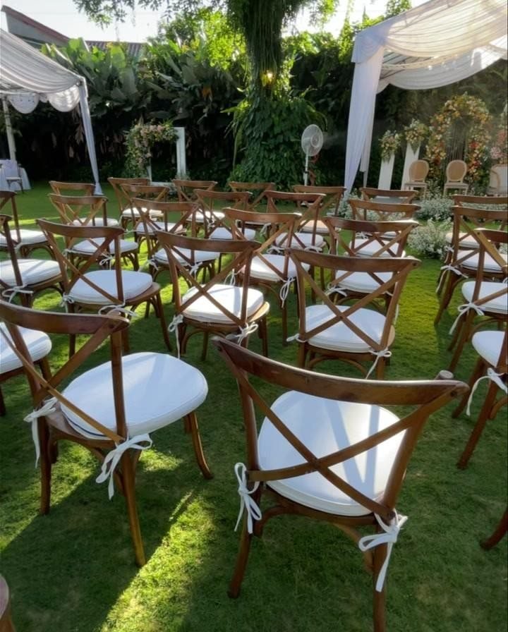 Wooden cross-back chairs with white cushions arranged on a green lawn for an outdoor wedding ceremony.