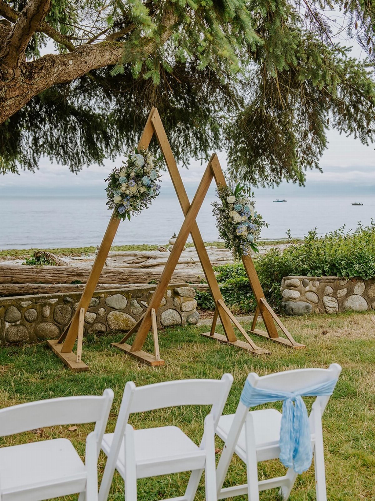 Two wooden triangle wedding arches decorated with blue and white flowers by the ocean.