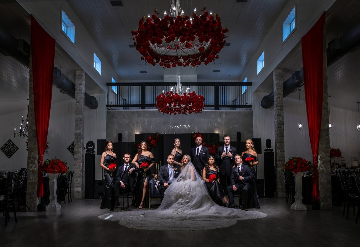 Wedding party in black and red attire posing in a decorated hall with red floral chandeliers.