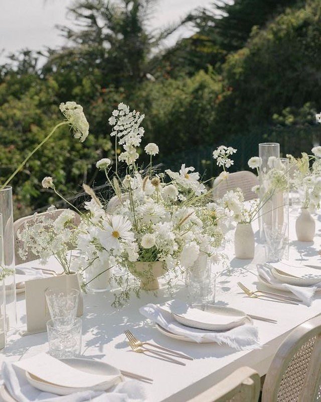 White table setting with floral centerpiece, gold cutlery, and white plates set outdoors near green trees.