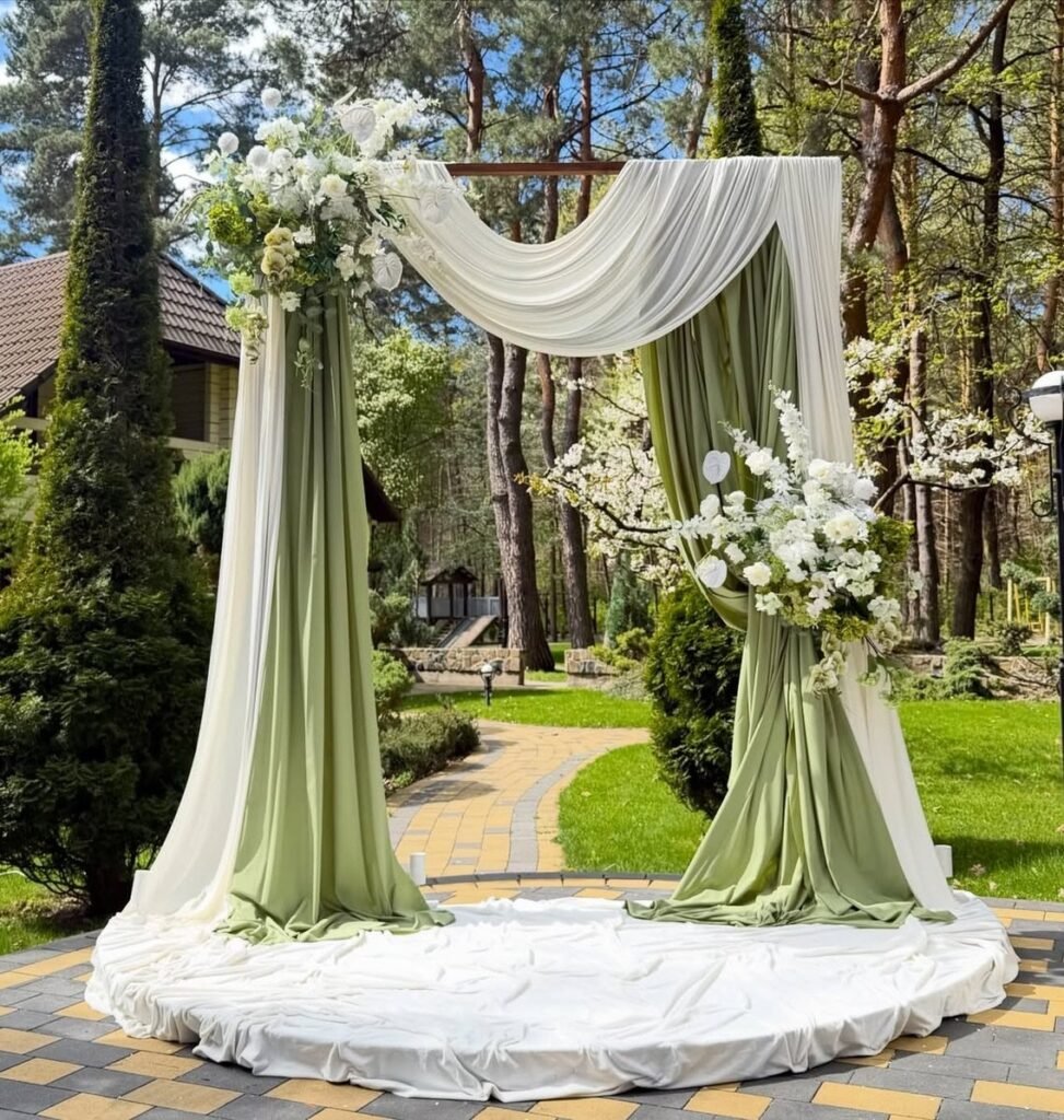 A wooden wedding arch draped in white and sage green fabric with white floral arrangements.