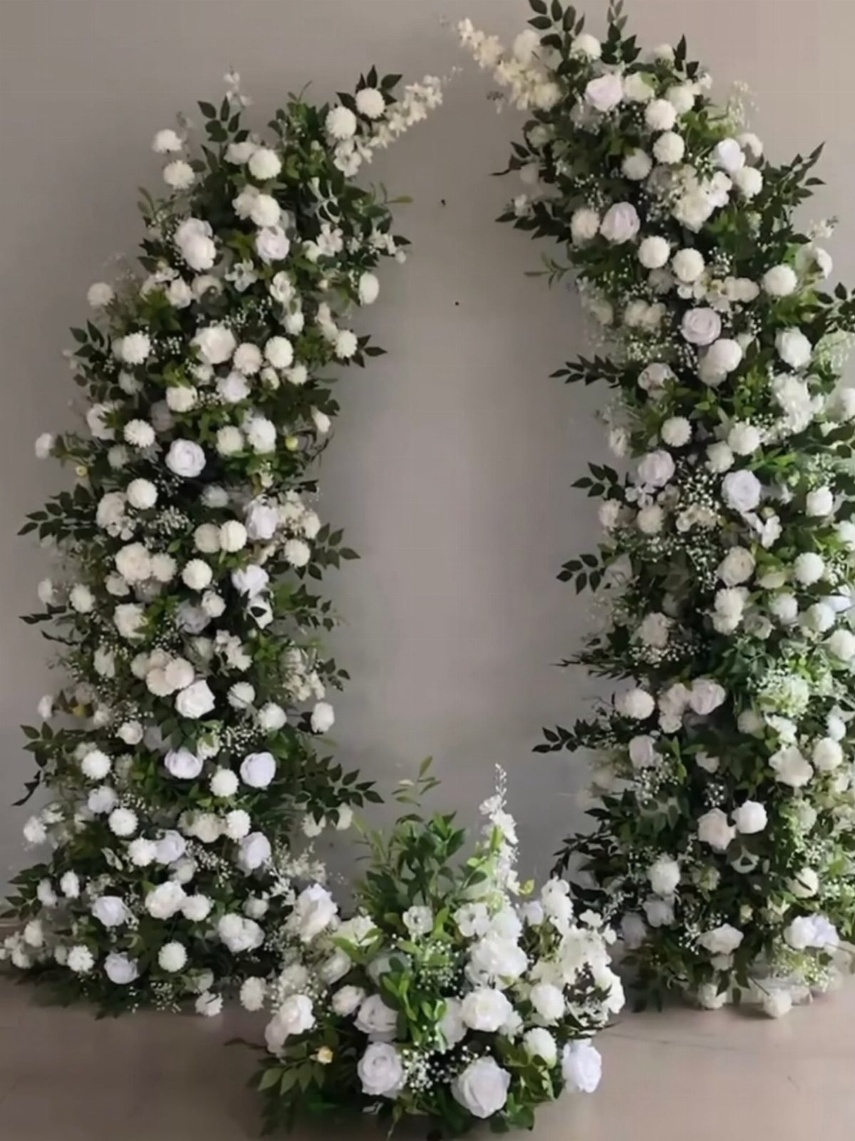 A large circular wreath made of white roses and green foliage against a grey wall.