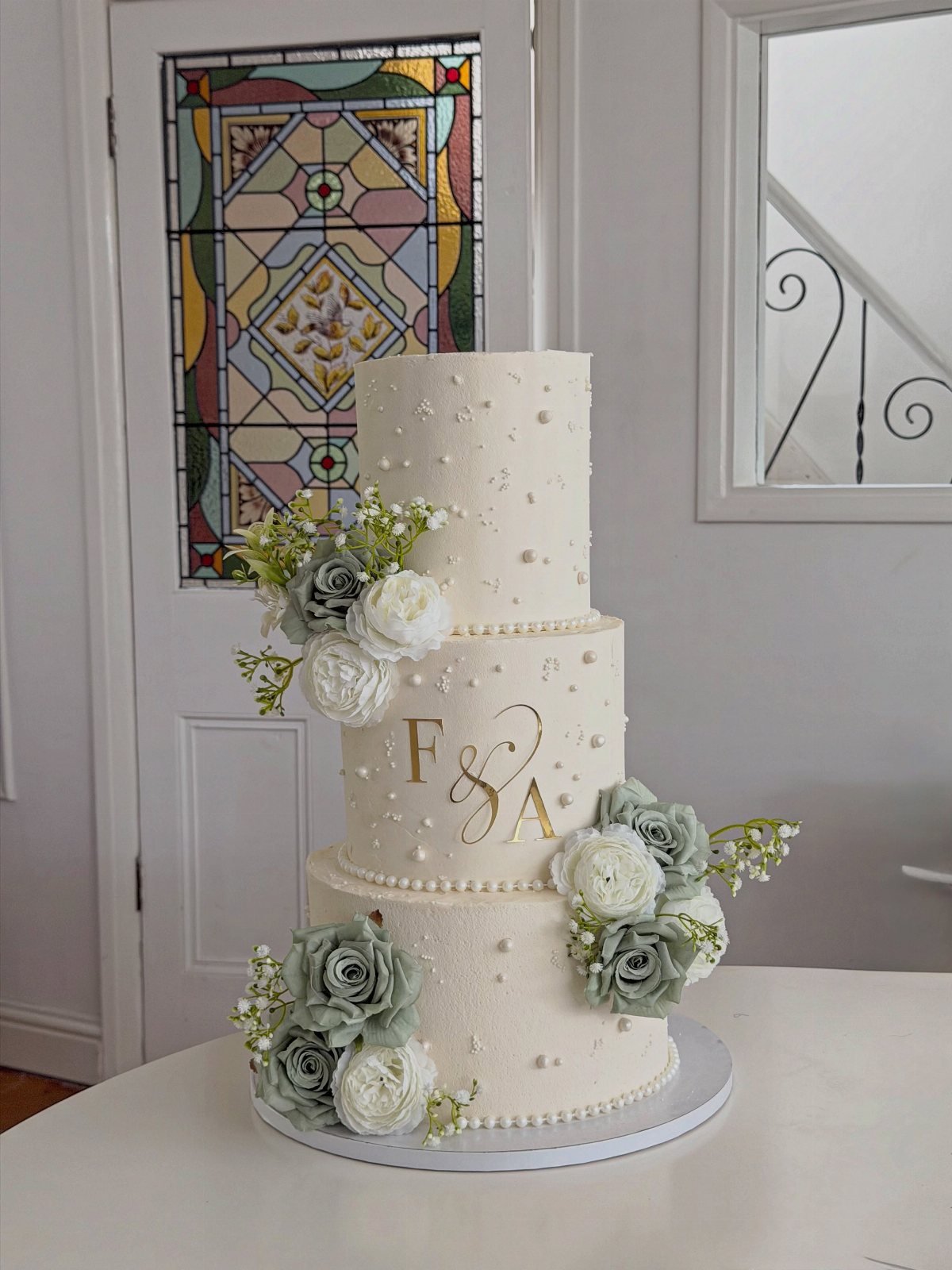 Three-tier white wedding cake decorated with pearls and sage green and white flowers.