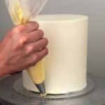 A hand piping yellow frosting onto the base of a tall white frosted cake.