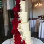 A four-tier white wedding cake with gold leaf and cascading deep red roses.