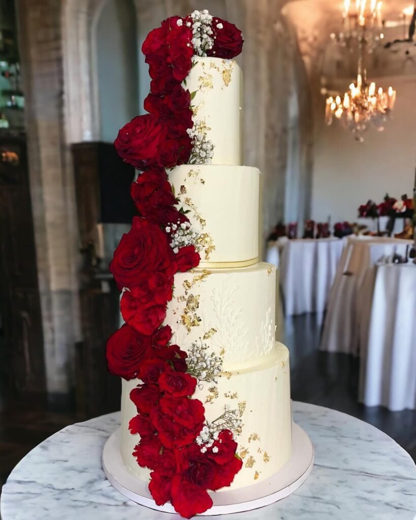 A four-tier white wedding cake with gold leaf and cascading deep red roses.