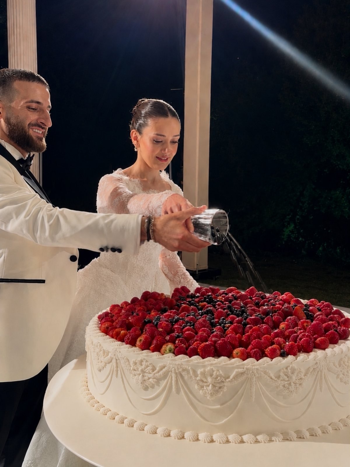 A white wedding cake topped with many fresh red strawberries and dark berries being served at night.
