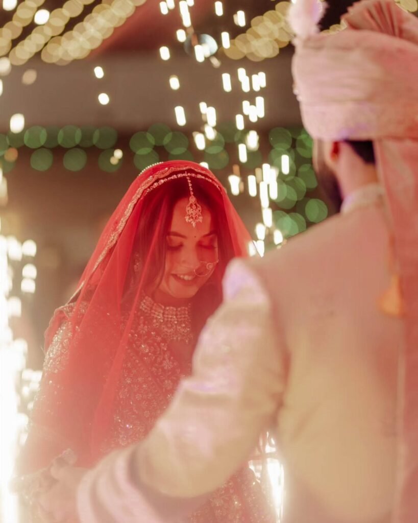 A smiling bride in a red veil and traditional jewelry stands near a groom in a pink turban.