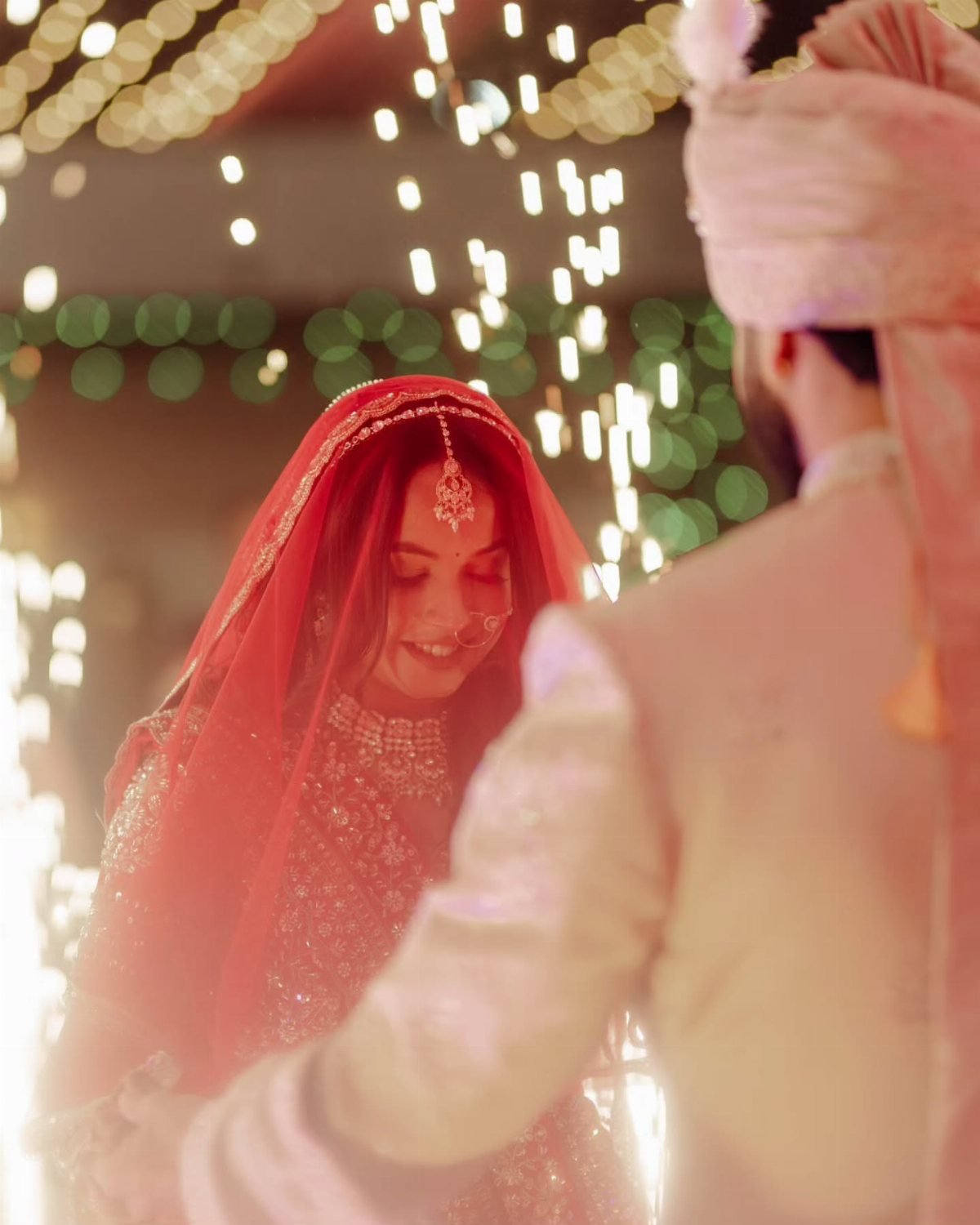 A smiling bride in a red veil and traditional jewelry stands near a groom in a pink turban.