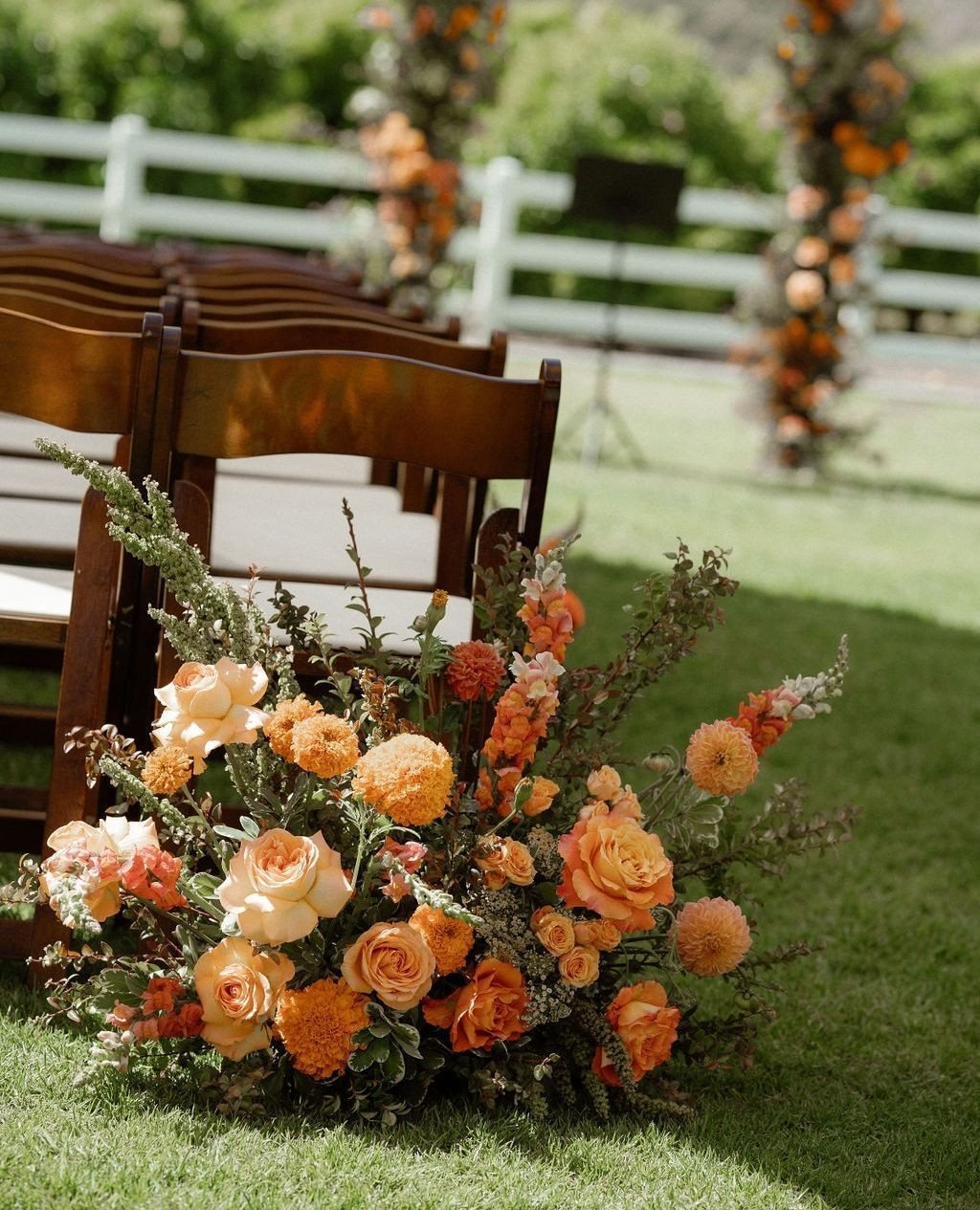 Orange roses and marigolds arranged in a floral bouquet on a green lawn near wooden chairs.