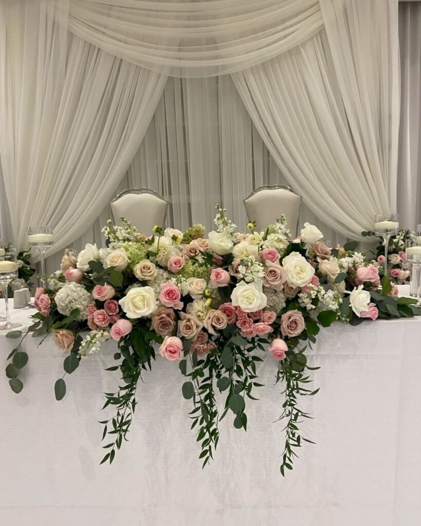 A lush floral arrangement of pink and white roses decorating a wedding head table with white draping.