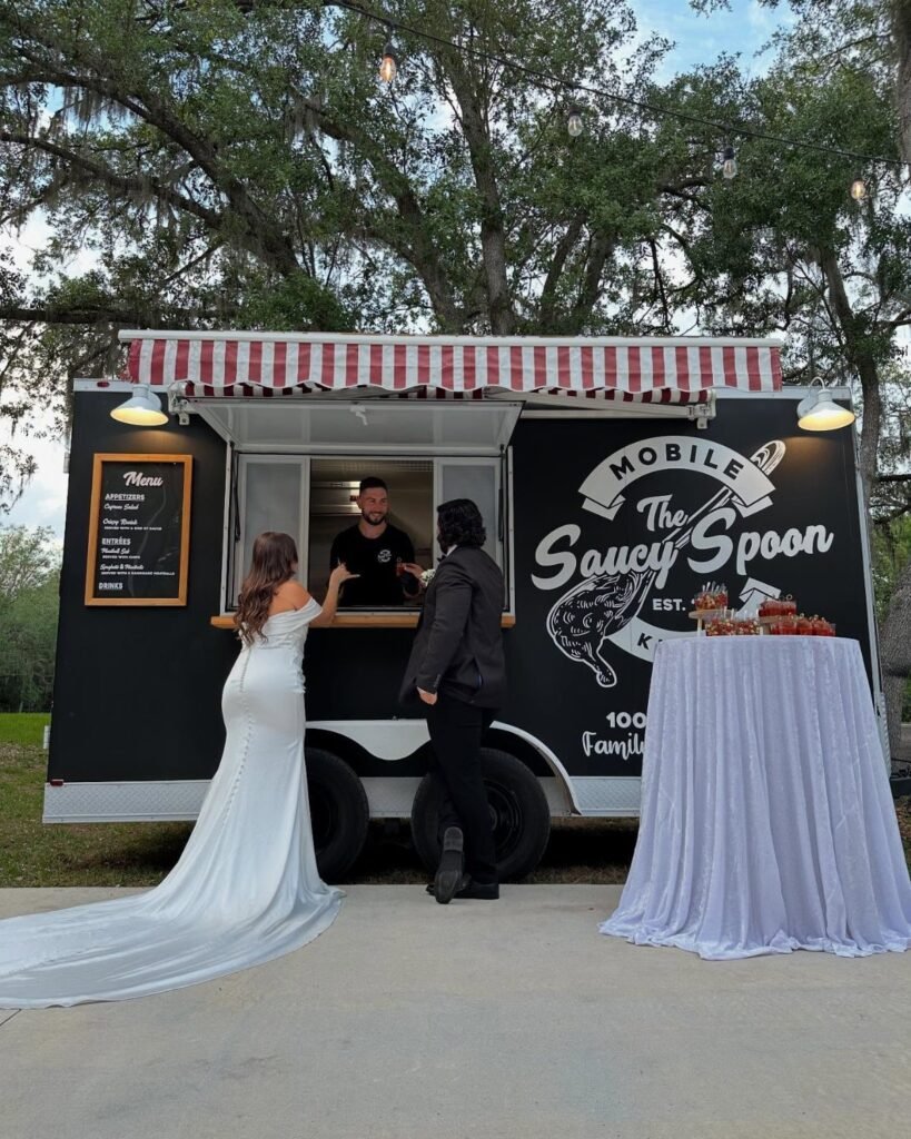 A bride and guest standing before a black mobile food truck under large trees.