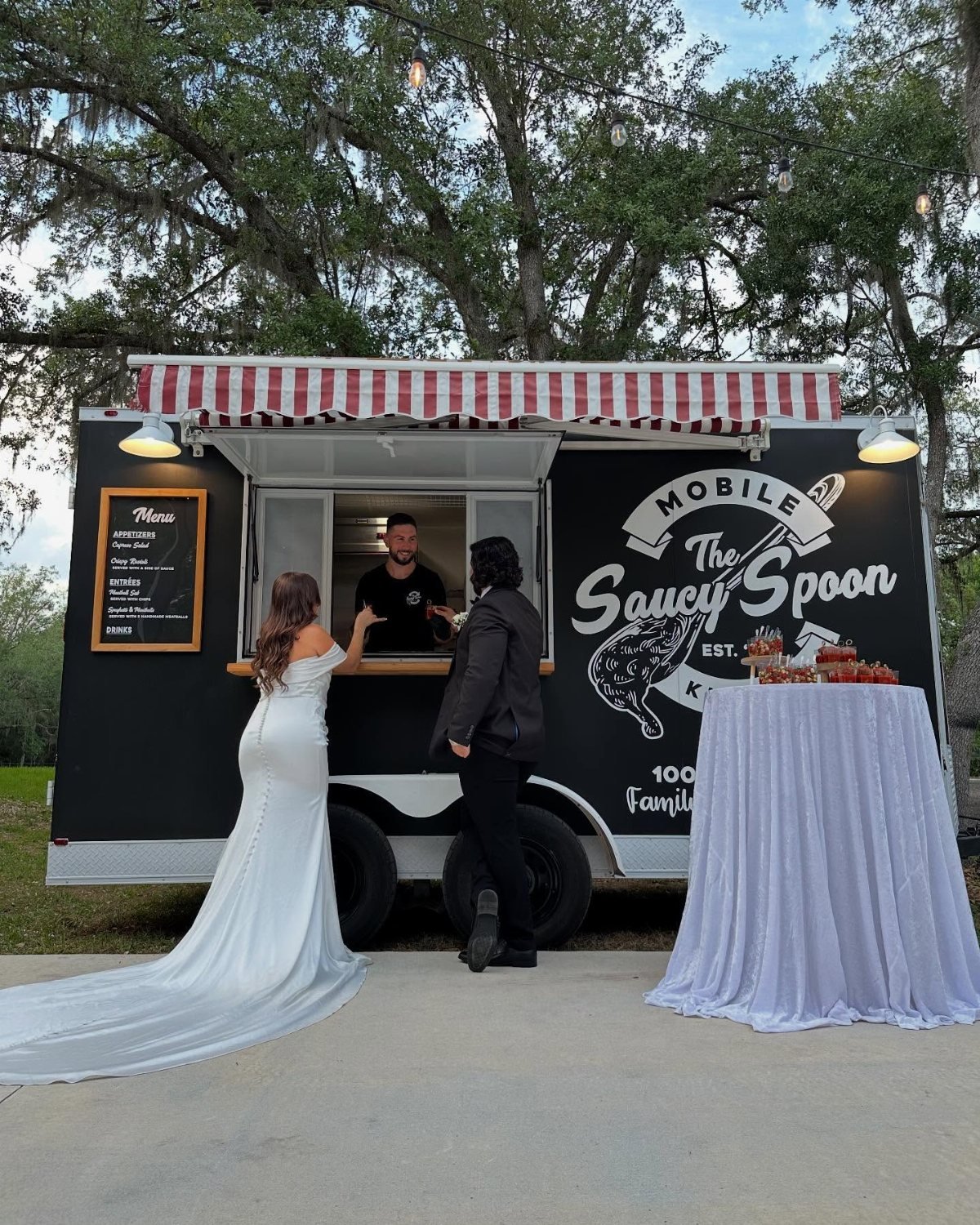 A bride and guest standing before a black mobile food truck under large trees.