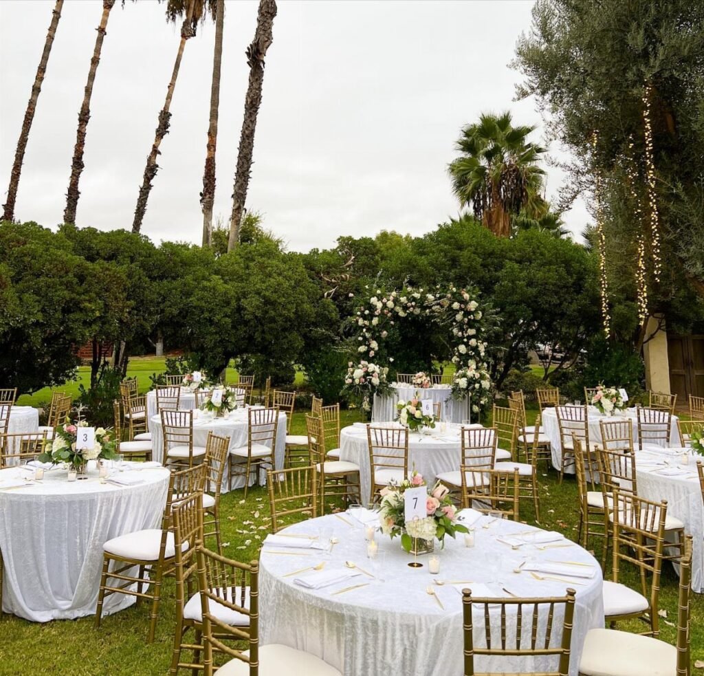 Wedding reception tables with white linens and gold chairs set on a green lawn under palm trees.