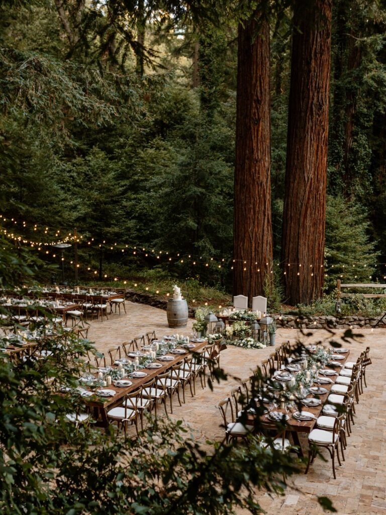 Long wooden dining tables set for a wedding reception under tall redwood trees and string lights.