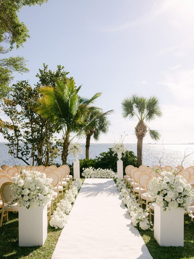 White wedding ceremony chairs and floral arrangements arranged on grass beside the ocean with palm trees.